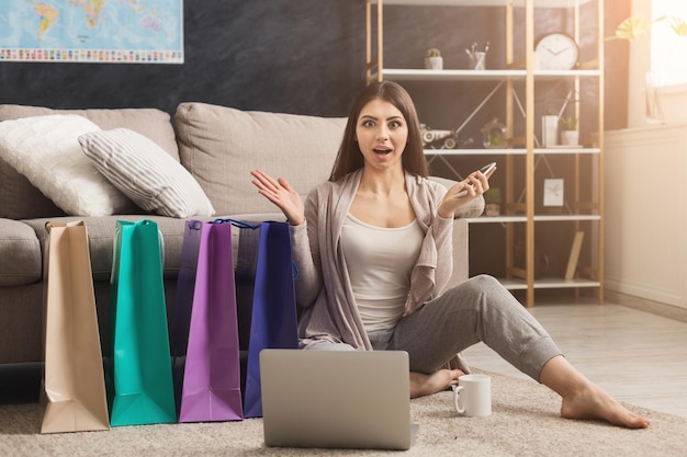 Happy surprised fashion woman buying online with laptop and credit card, sitting on floor with colorful shopping bags, copy space