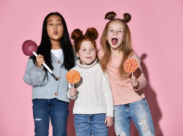 Happy surprised girls screaming and laughing while posing with huge lollipop and spiral sweet candies studio portrait isolated on pink background Funny crazy facial expression and sugar dependence