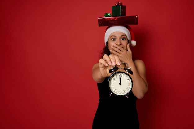 Happy surprised gorgeous woman in black dress and Santa hat, with gift boxes on head, covers her mouth showing an alarm clock with time around 12 o'clock at night, looking at camera. Christmas concept