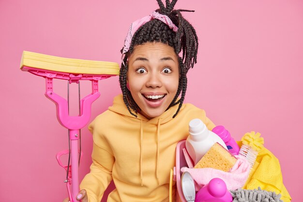 Happy surprised housewife tries to keep house clean gets challenging tasks poses with laundry basket and mop