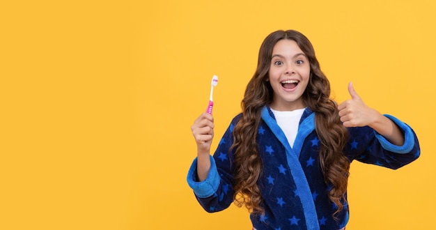 Happy surprised kid girl in sleepwear show thumb up and toothbrush copy space hygiene