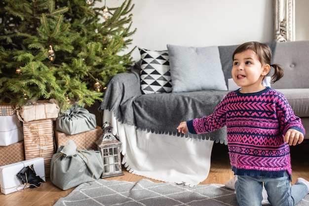 Happy surprised toddler girl to see a lot holiday gifts under Christmas tree Merry Christmas and Happy New Year Cute little child girl choose the present Beautiful girl and Christmas tree indoors