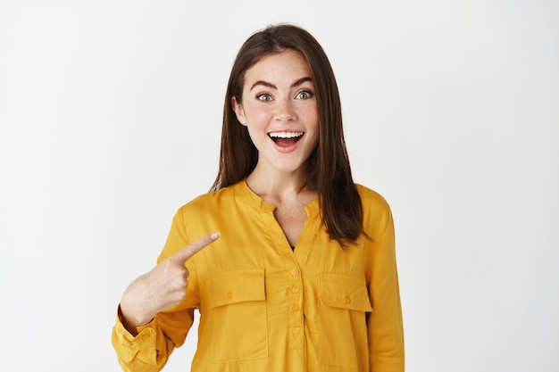 Happy and surprised woman pointing at herself, looking amazed at camera and smiling, being chosen, standing over white wall