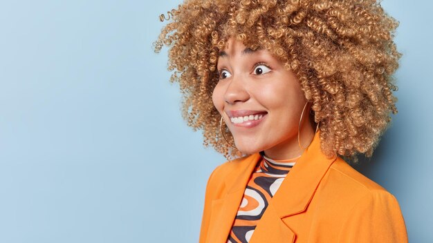Happy surprised woman with curly bushy hair bites lips feels\
amazed cannot believe in amazing news dressed in elegant orange\
jacket isolated over blue background blank space human\
reactions