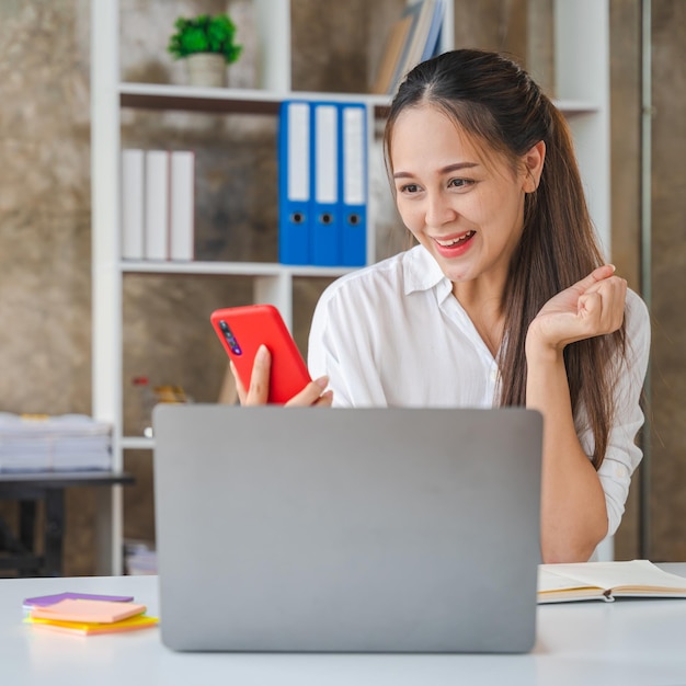 Счастливая удивленная молодая женщина работает с ноутбуком Счастливая взволнованная азиатская деловая женщина в офисе работает с мобильным телефономДа, отличная работа