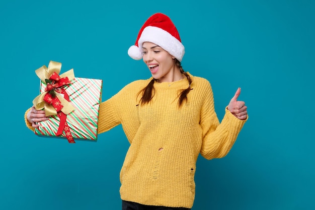 Happy surprised young woman in santa claus hat holding new year gift laughing and showing thumb up gesture on blue isolated background