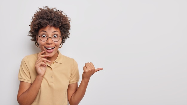 Photo happy surprised young woman with curly hair points thumb away shows amazing news demonstrates promotion dressed in casual wear isolated over white background. people and advertisement concept