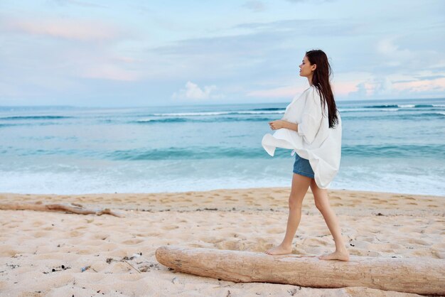 写真 白い水着シャツとジーンズのショートパンツを着た幸せな日焼けしたスリムな女性は、夕日の光を泳いだ後、濡れた髪で海の砂の上のビーチを歩きます