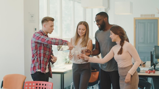 Photo happy team drinking alcohol in workplace young man opening bottle of champagne