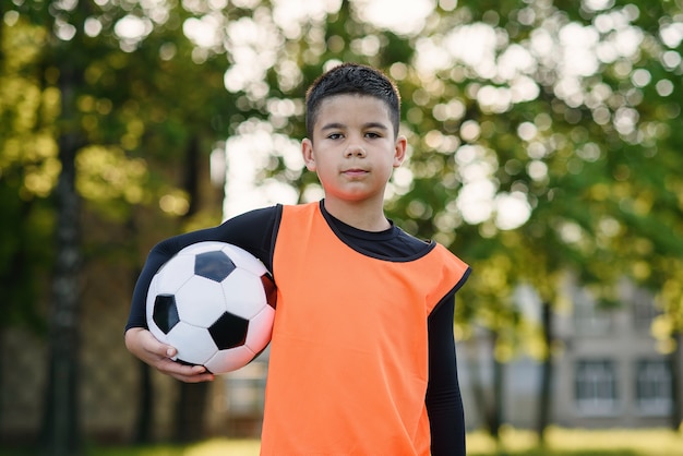 サッカーの制服を着た幸せな10代の少年は、朝のトレーニング後にボールを保持しています。