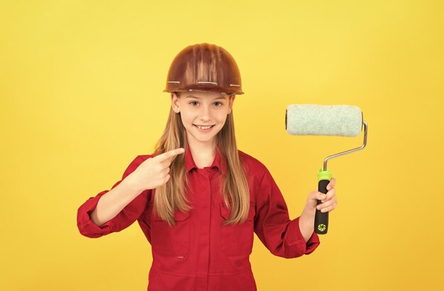 Happy teen child in builder hard hat with paint roller on yellow wall point finger