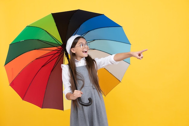Happy teen girl in glasses and beret under colorful umbrella for rain protection in autumn season pointing finger on copy space advertising