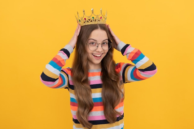 Happy teen girl in glasses wear princess crown on yellow background, prom