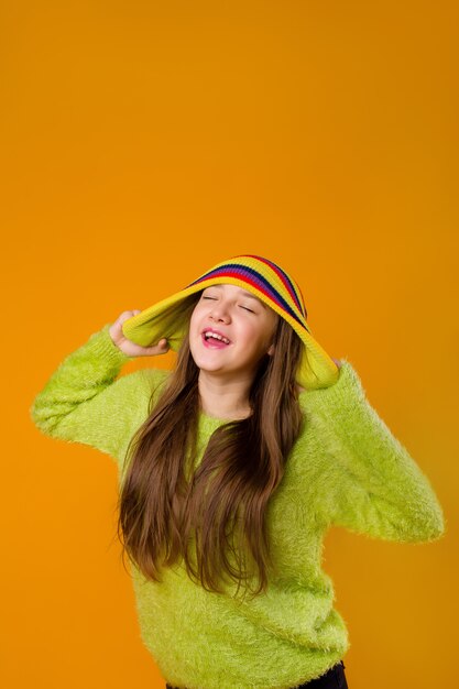 Happy teen girl in green sweater and multi-colored knitted hat on a yellow background