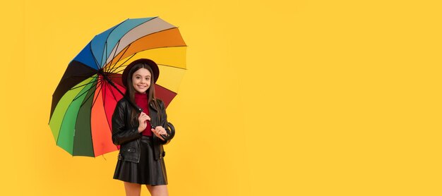 Happy teen girl in hat and leather clothes under rainbow umbrella autumn rain Child with autumn umbrella rainy weather horizontal poster banner with copy space