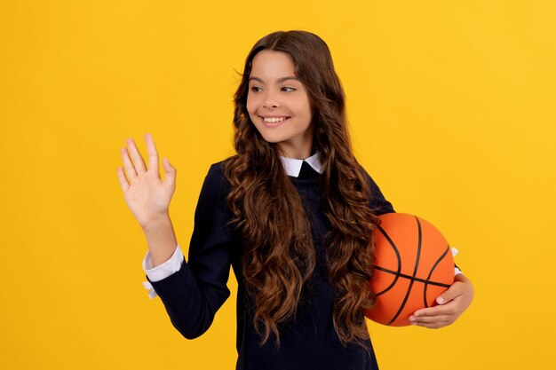 Happy teen girl hold basketball ball waving hello on yellow background sport school