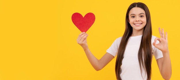 Ragazza teenager felice che tiene il cuore di san valentino che mostra un gesto ok su sfondo giallo amore ragazza del bambino