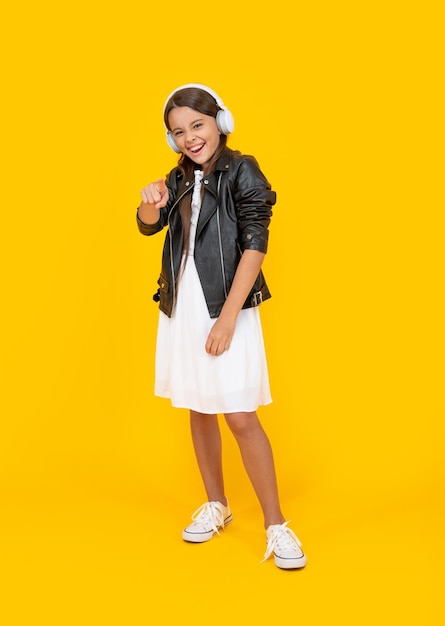 Happy teen girl listen music in headphones on yellow background pointing finger