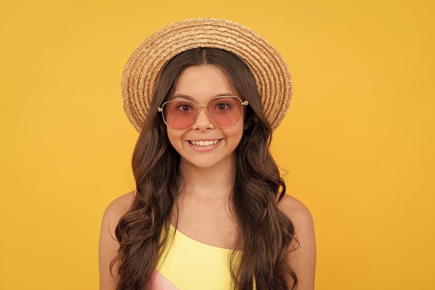 Happy teen girl in summer straw hat and glasses has curly hair on yellow background portrait