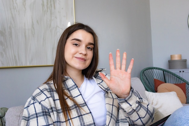 Happy teen girl waving hand video conference calling on tablet computer sit on coach