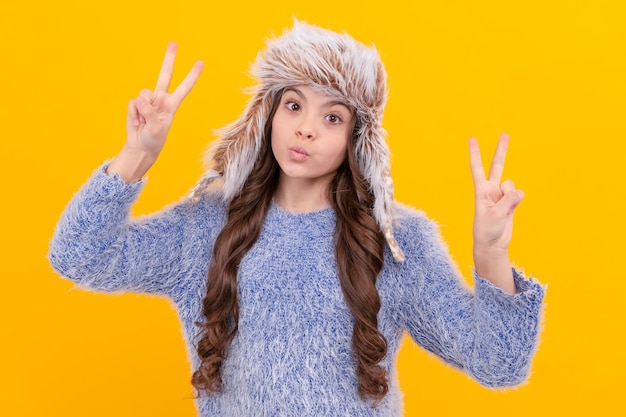 Happy teen girl wear earflap hat showing peace gesture on yellow background, winter