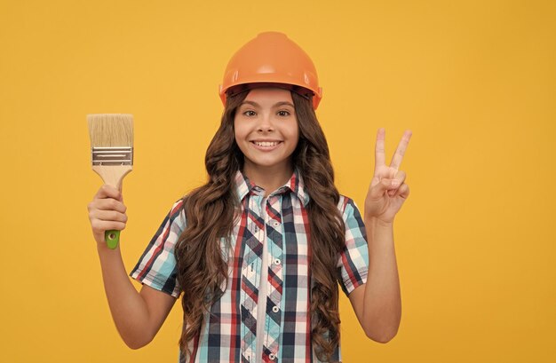 Happy teen girl with curly hair in construction helmet hold painting brush show peace gesture decoration