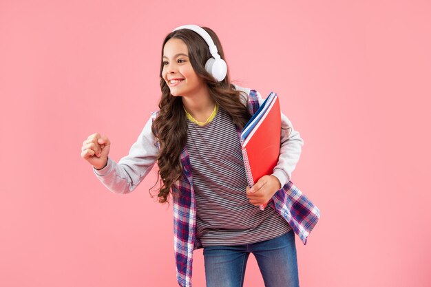 Ragazza teenager felice con la cartella di lavoro che indossa le cuffie su sfondo rosa, scuola
