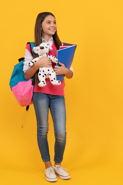 Счастливая школьница-подросток с рюкзаком и тетрадью держит игрушку на желтом фоне