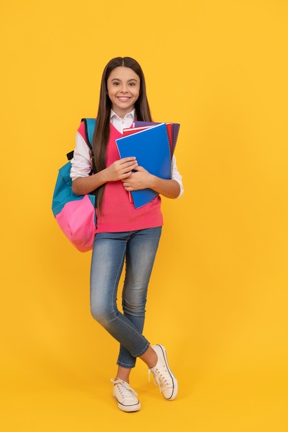 Happy teen school girl with backpack and copybook on yellow background education