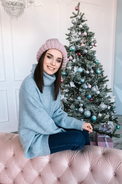 Donna teenager felice che si siede in un sofà vicino all'albero di natale. vestito con un maglione e un cappello di lana lavorati a maglia blu. una piacevole sorpresa. regali di capodanno.