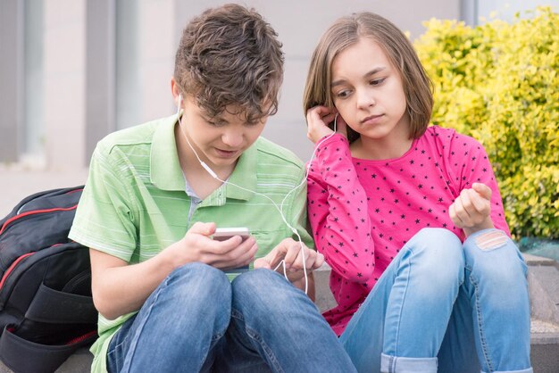 Photo happy teenage boy and girl with headphones are using gadget talking and smiling while sitting on the stairs outdoors young sister and brother teens playing on mobile phone and listening to music