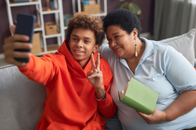 Happy teenage boy and his mature mother with book looking at smartphone camera