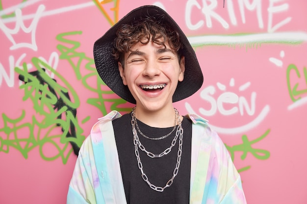 Happy teenage boy with braces on teeth wears stylish clothes with chains around neck has fun with friends poses against colorful graffiti wall as example of modern art