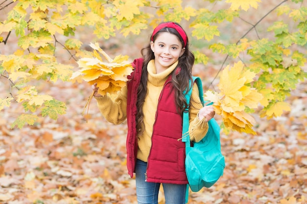 Фото Счастливый ребенок-подросток, наслаждаясь осенним лесом с красивой сезонной природой, несет школьную сумку и держит желтые опавшие кленовые листья обратно в школу