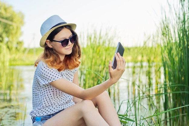 Happy teenage girl in hat with smartphone on wooden pier of lake with reeds, summer vacation in nature, girl communicates online with friends, blogs in social networks