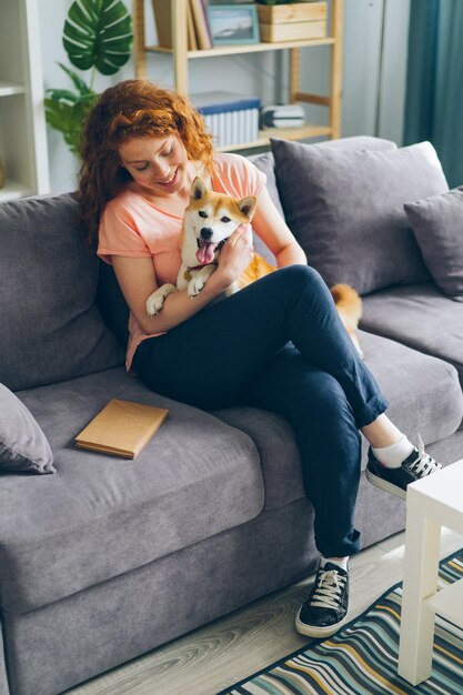 Happy teenage girl hugging dog sitting on sofa in modern apartment smiling