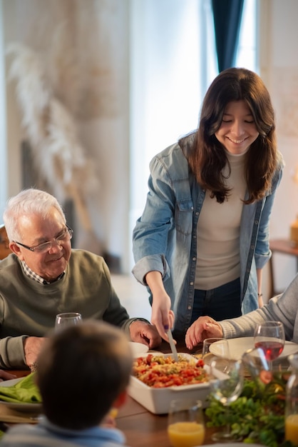 Happy teenage girl serving pasta at family dinner. Multigenerational family sitting at table during party. Family party concept