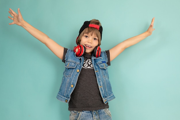 Ragazzo felice dell'adolescente in pantaloncini di jeans e giacca, maglietta nera, berretto nero, con sorrisi di eaphones rossi e mani in alto