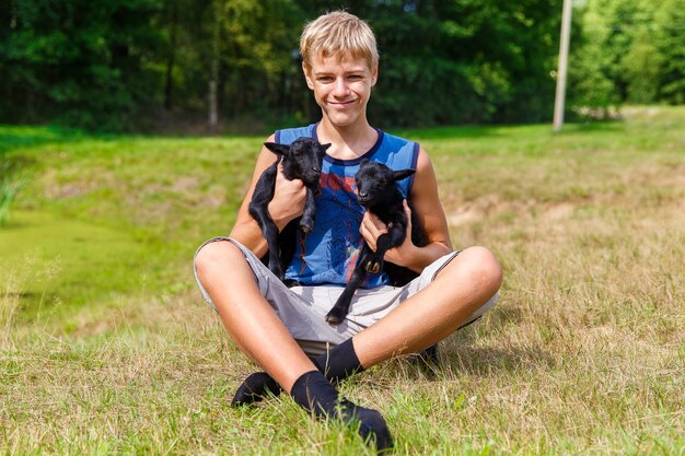 Happy teenager boy holding little black baby goats in the meadow.