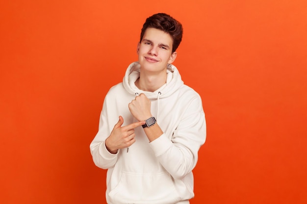 Happy teenager in casual style hoodie showing smartwatch on his wrist with smile on face, satisfied with gadget, contactless payments. indoor studio shot isolated on orange background