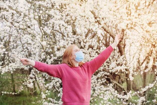 春の花の咲く庭園で医療マスクで幸せなティーンエイジャーの女の子。社会的距離の概念とコロナウイルスの予防。