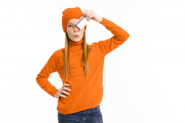 Happy teenager girl with red hair, orange hoody and hat holds a credit card isolated on white background