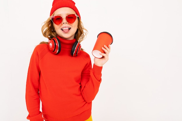 Happy teenager girl with red hair, red glasses smiles and drinks coffee isolated on white