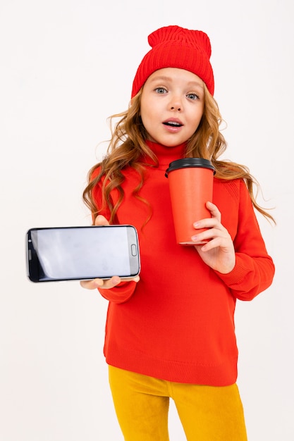 Happy teenager girl with red hair, red hat, hoody and yellow trousers smiles, drinks coffee and play phone games isolated on white