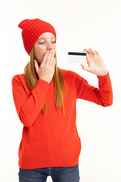 Happy teenager girl with red hair, red hoody and hat with card isolated on white