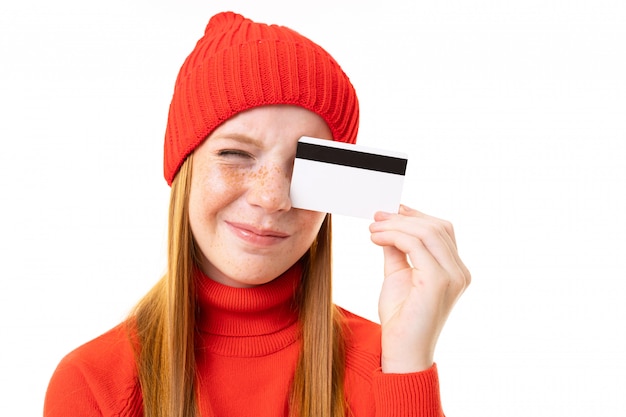 Happy teenager girl with red hair, red hoody and hat with credit card isolated on white