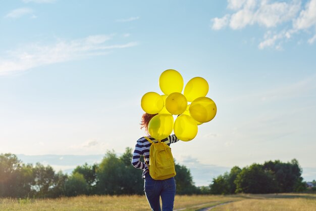 Happy teenager girl with yellow balloons and backpack running\
and jumping along country road in summer meadow. freedom, life,\
joy, holiday concept