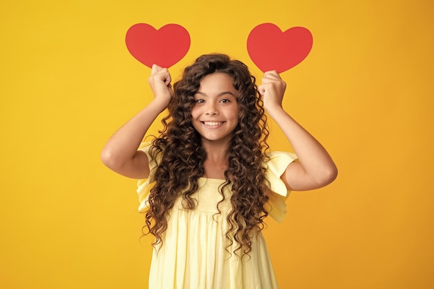 Happy teenager portrait Cheerful lovely romantic teen girl hold red heart symbol of love for valentines day isolated on yellow background Smiling girl