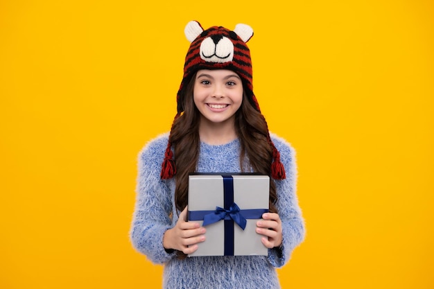 Happy teenager positive and smiling emotions of teen girl teenager child in funny winter hat holding gift box on yellow isolated background gift for kids birthday christmas or new year