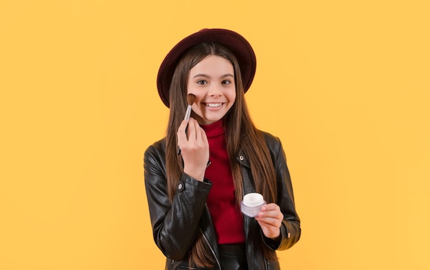 Happy ten girl in hat apply powder with makeup brush on yellow background beauty